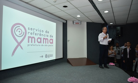 Em cima do palco, o secretário municipal da saúde; Ao fundo, uma tela com o logo do Serviço de Referência de mama.